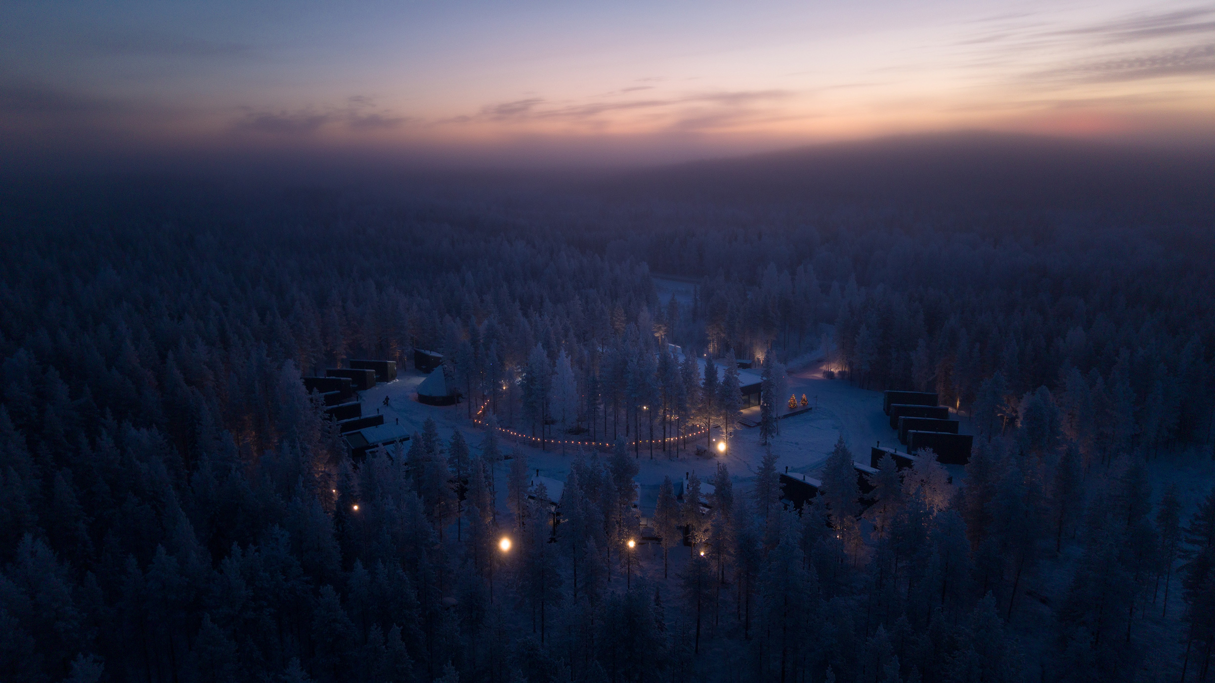Luchtfoto van Halo Igloos Rovaniemi in de schemering, met verlichte glazen iglo’s in het sneeuwbos